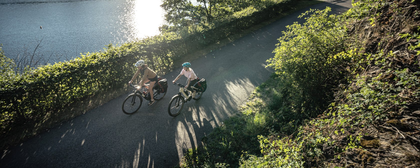 Zwei Radfahrer fahren bei Sonnenschein entlang eines Sees, umgeben von grüner Natur. Die Sonne spiegelt sich im Wasser., © Eifel Tourismus GmbH, Dennis Stratmann Zwei Radfahrer fahren bei Sonnenschein entlang eines Sees, umgeben von grüner Natur. Die Sonne spiegelt sich im Wasser., © Eifel Tourismus GmbH, Dennis Stratmann