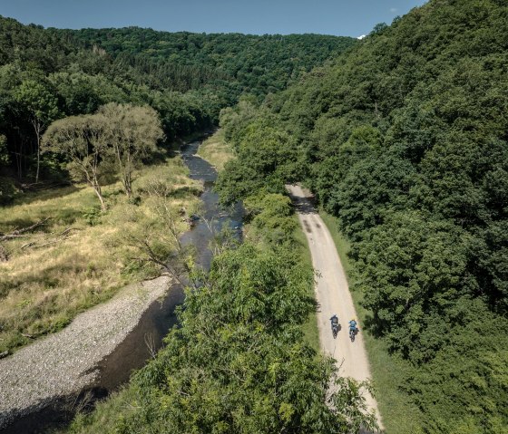 Two cyclists ride on a path along a river, surrounded by a dense, green forest landscape under a clear blue sky., © Eifel Tourismus GmbH, Dennis Stratmann Two cyclists ride on a path along a river, surrounded by a dense, green forest landscape under a clear blue sky., © Eifel Tourismus GmbH, Dennis Stratmann