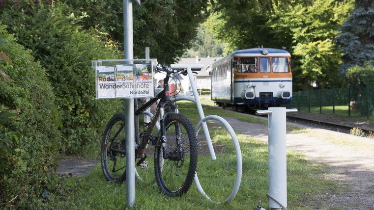 Ein Fahrrad steht an einem Parkplatz mit einem Schild für den Radweg. Im Hintergrund fährt ein Zug vorbei.