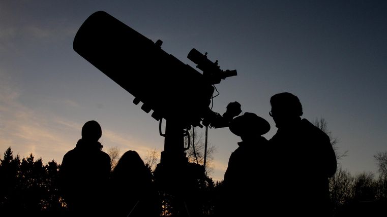 Silhouettes of people looking at the evening sky through a telescope. The sky is dark in the background, allowing for a clear view of the stars.