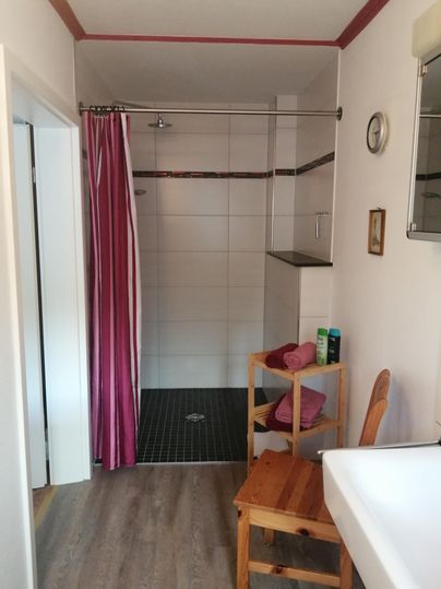 Ein modernes Badezimmer mit einer Dusche, die von einer gestreiften Vorhang abgetrennt ist. Auf einem Holzstuhl liegen Handtücher, und ein Waschbecken ist im Vordergrund zu sehen.