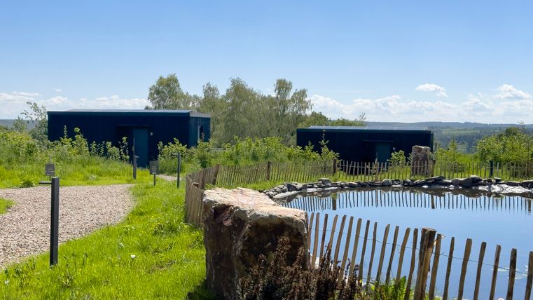 Zwei moderne Wohncontainer stehen in einer grünen Landschaft neben einem kleinen Teich. Der Himmel ist klar und die Umgebung ist ruhig und naturnah.