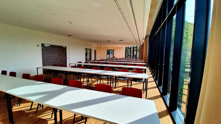 Ein heller Seminarraum mit langen Tischen und roten Stühlen. Große Fenster bieten viel Tageslicht und einen Blick nach draußen.