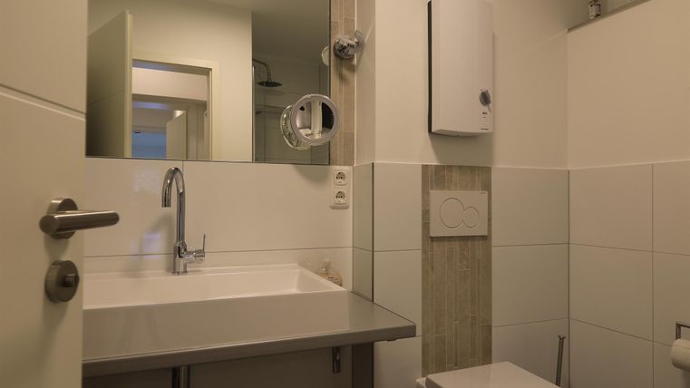 Ein modernes Badezimmer mit einem Waschtisch, einem Spiegel und einer Toilette. Die Wände sind in neutralen Farben gehalten, und es gibt eine Dusche außerhalb des Sichtfelds.