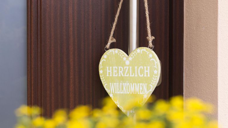 Ein herzliches Willkommensschild in Herzform hängt an einer Tür. Im Vordergrund sind gelbe Blumen zu sehen.