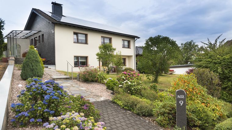Ein modernes Haus mit einem schönen Garten und bunten Blumen. Der Weg zum Haus ist gepflastert und es ist eine ruhige Atmosphäre zu sehen.