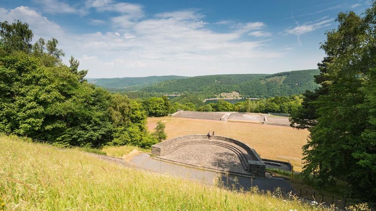 Eine Aussicht auf ein Freiluft-Amphitheater, umgeben von Bäumen und sanften Hügeln. Der Himmel ist klar und blau, ideal für Veranstaltungen im Freien.