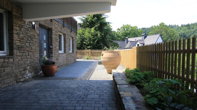 Ein gepflasterter Weg vor einem modernen Gebäude mit Pflanzen. Im Hintergrund sind grüne Bäume und eine Holzmauer zu sehen.