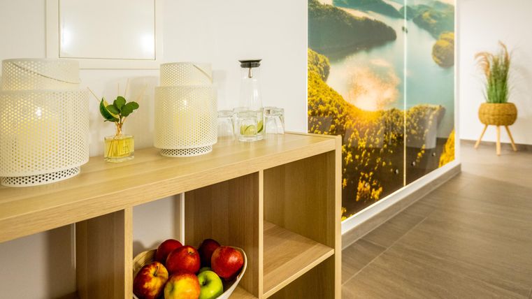 Ein heller Raum mit einem modernen Holzregal und einer Schüssel voll Äpfeln. Im Hintergrund ist eine Wand mit einer Landschaftsdarstellung zu sehen.