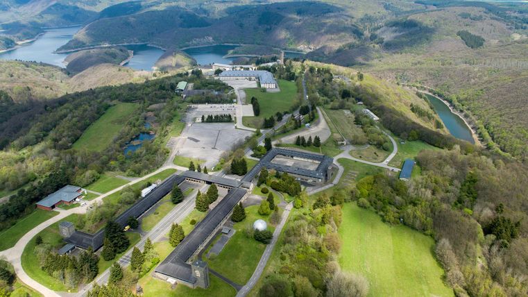 Eine Luftaufnahme von einer grünen Landschaft mit Gebäuden und Straßen. Im Hintergrund sind Berge und ein Gewässer zu sehen.
