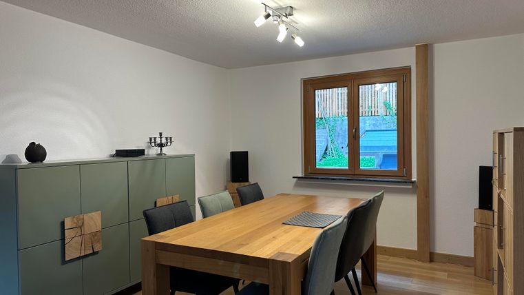 Ein modernes Esszimmer mit einem großen Holztisch und Stühlen. Ein Fenster lässt natürliches Licht herein, während eine grüne Aussicht sichtbar ist.