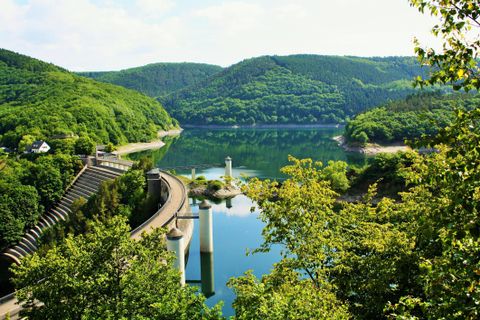 <p>Man sieht die Staumauer von oben. Ringsherum Bäume und glitzerndes Wasser.</p>