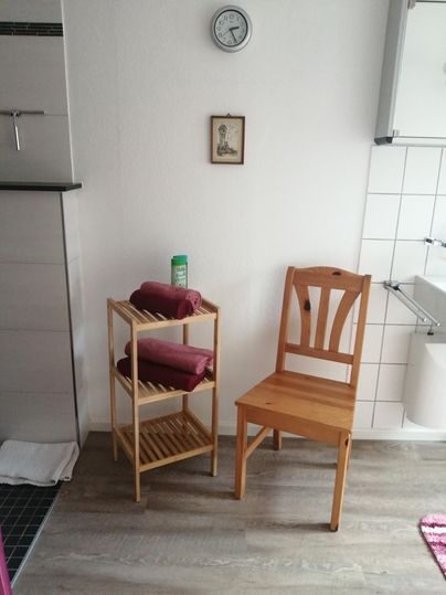 Ein einfaches Badezimmer mit einem Holzstuhl und einem Regal. Das Regal ist mit Handtüchern und einer Flasche dekoriert.