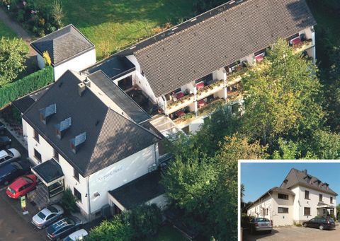 Ein Luftbild eines mehrstöckigen Gebäudes mit Balkonen und begrünten Bereichen. Im Vordergrund sind mehrere Parkplätze und Autos zu sehen.