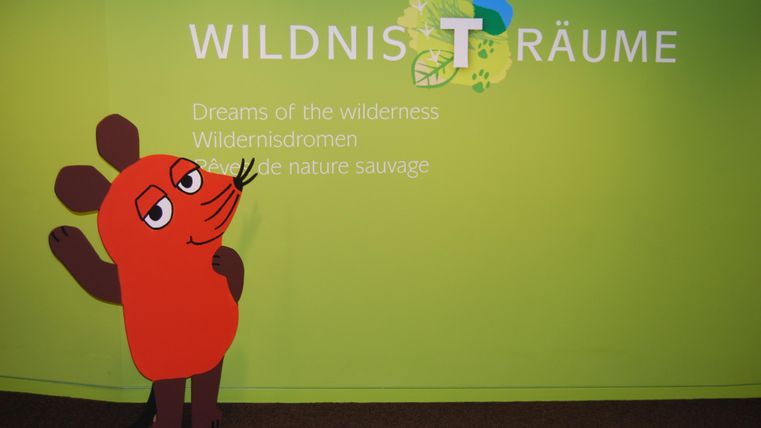 Das Bild zeigt die Maus von Die Sendung mit der Maus vor dem Wildnis-Träume Schriftzug.