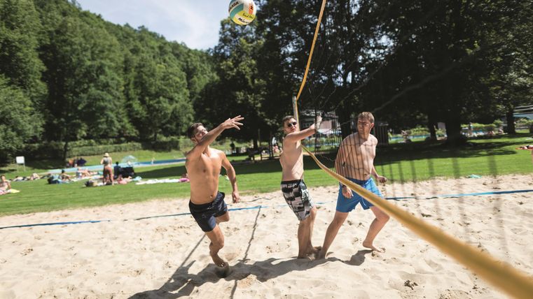 Drie mannen spelen beachvolleybal op een zonnige dag. De omgeving is groen en uitnodigend, met veel mensen op de achtergrond.