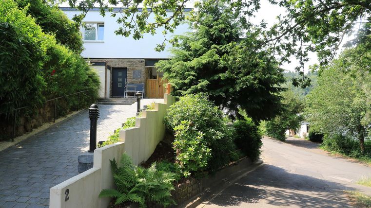 Ein modernes Haus mit einem gepflegten Vorgarten und viel Grün. Der Weg zum Haus ist gepflastert und es gibt Bäume und Sträucher in der Umgebung.