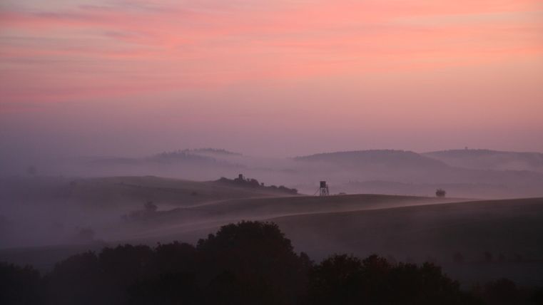 Eine sanfte Landschaft mit sanften Hügeln und Nebel. Der Himmel zeigt ein schönes Farbenspiel bei Sonnenaufgang.