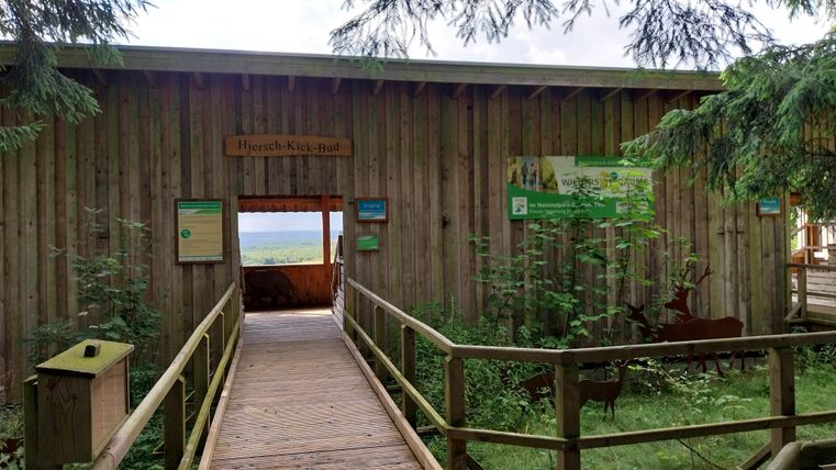 Ein Holzgebäude mit einem breiten Holzsteg, der dorthin führt. Umgeben von Bäumen und einer grünen Landschaft.