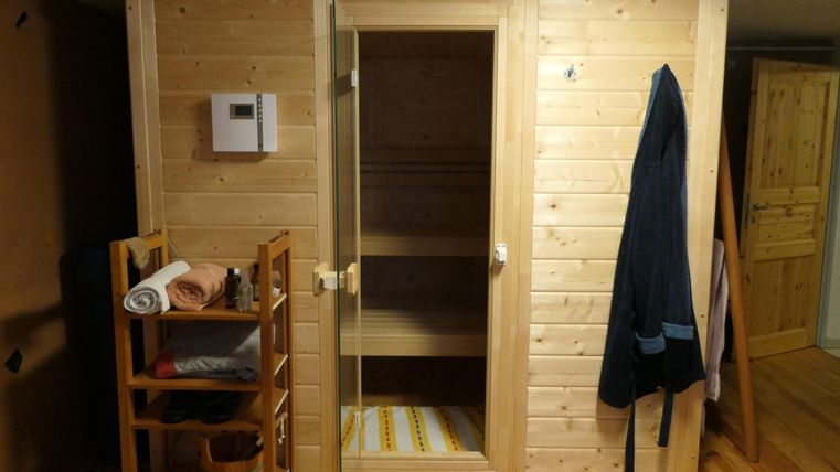 Ein gemütlicher Saunabereich aus hellem Holz mit einer Glastür. Neben der Sauna steht ein Regal mit Handtüchern und Sauna-Zubehör.
