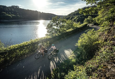 Zwei Radfahrer fahren bei Sonnenschein entlang eines Sees, umgeben von gr&uuml;ner Natur. Die Sonne spiegelt sich im Wasser., &copy; Eifel Tourismus GmbH, Dennis Stratmann
