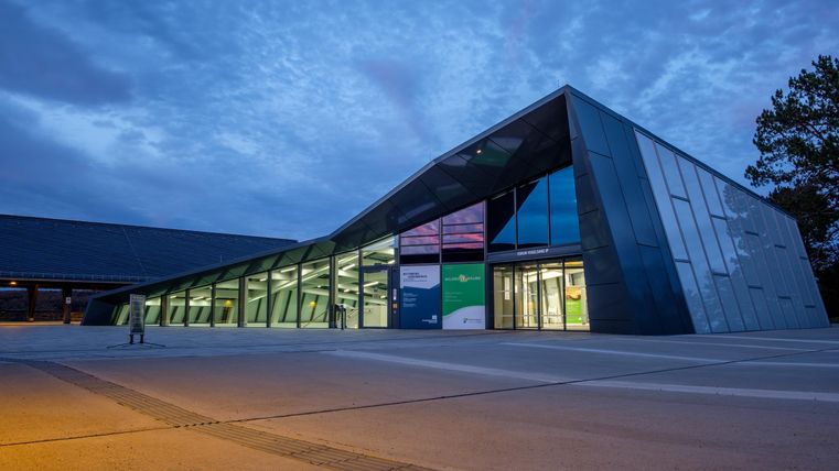 Ein modernes Gebäude mit einer markanten, schrägen Architektur. Es ist zum Abend hin beleuchtet, und der Himmel ist teilweise bewölkt.