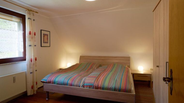 Ein einfaches Schlafzimmer mit einem Doppelbett und bunten Streifenbettwäsche. Die Wände sind hell, und es gibt ein Fenster mit Vorhängen.