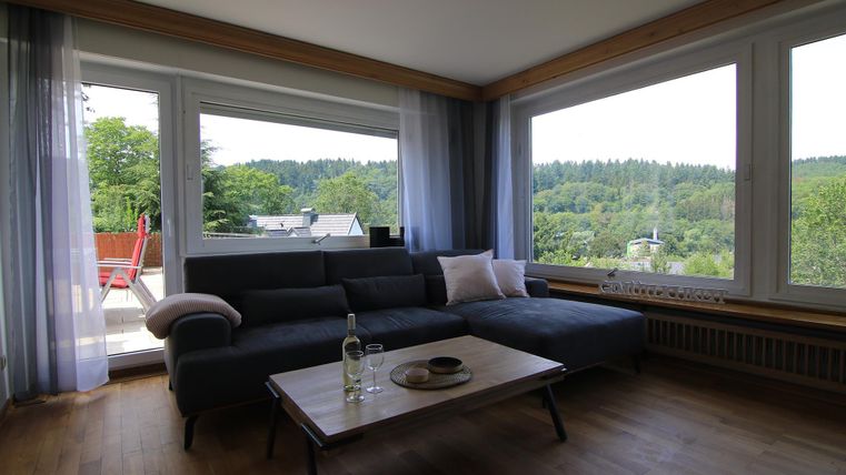 Ein helles Wohnzimmer mit großen Fenstern und Blick auf die grüne Landschaft. Ein gemütliches Sofa und ein niedriger Tisch sorgen für eine einladende Atmosphäre.