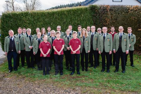 Eine Gruppe von Menschen steht im Freien, umgeben von grünen Hecken und einem Gebäude im Hintergrund. Die Mitglieder tragen einheitliche Jacken und Hemden in verschiedenen Farben.