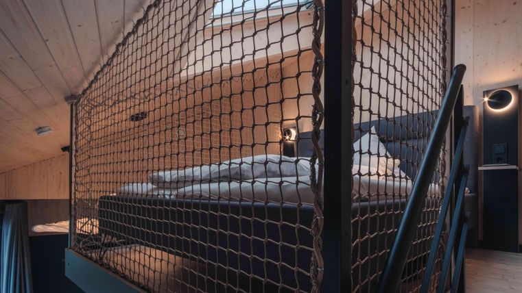 Ein modernes Schlafzimmer mit einem Podestbett, das durch ein Netz gesichert ist. Die Wände sind aus hellem Holz, und es gibt sanfte Beleuchtung für eine gemütliche Atmosphäre.