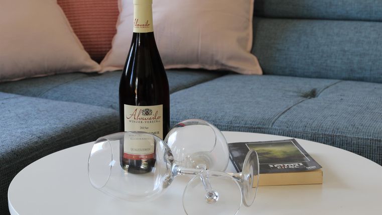 Eine Flasche Rotwein steht auf einem weißen Tisch. Daneben liegen zwei umgekehrte Weingläser und ein Buch.