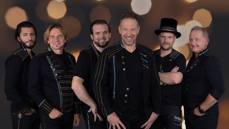 Eine Musikgruppe steht zusammen und lächelt in die Kamera. Im Hintergrund sind helle Lichtpunkte zu sehen.