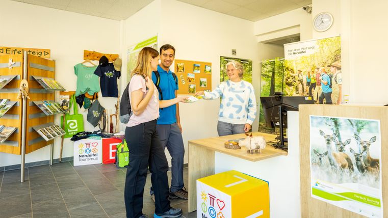 Une réception accueillante avec deux clients et une employée. Il y a des brochures et des articles de marchandise pour les randonneurs.