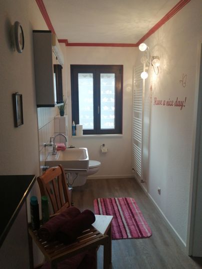 Ein helles Badezimmer mit einem Waschbecken, einem Fenster und einer Dusche. An der Wand steht der Spruch "Have a nice day!".