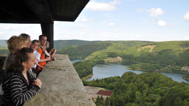 Eine Gruppe von Menschen steht an einer Aussichtsplattform und blickt auf eine malerische Landschaft mit einem See und grünen Hügeln. Der Himmel ist blau mit einigen Wolken.
