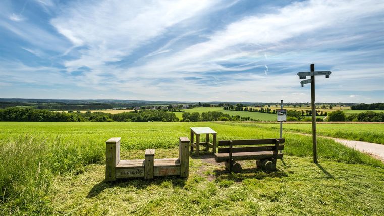 Eine ruhige Aussicht mit einem Sitzplatz aus Stein und Holz. Im Hintergrund erstrecken sich grüne Felder unter einem blauen Himmel.