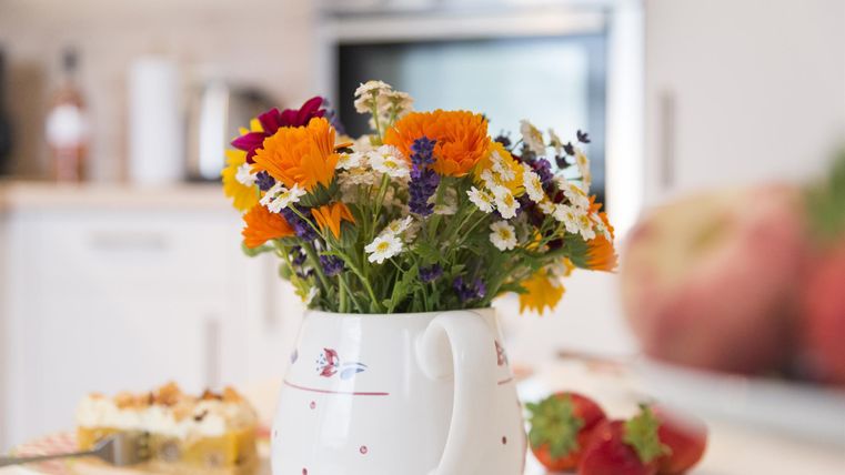 Ein bunter Blumenstrauß in einer weißen Kanne steht auf dem Tisch. Im Hintergrund sind Obst und ein Stück Kuchen sichtbar.