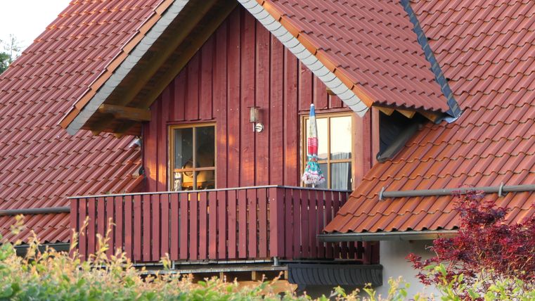 Ein charmantes Holzhaus mit rotem Dach und einem kleinen Balkon. Pflanzen umgeben das Haus und verleihen ihm eine einladende Atmosphäre.