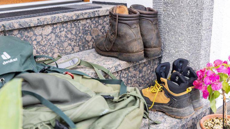 Ein paar robuste Stiefel stehen auf einer Treppe neben einem Rucksack. Im Vordergrund sind bunte Blumen zu sehen.