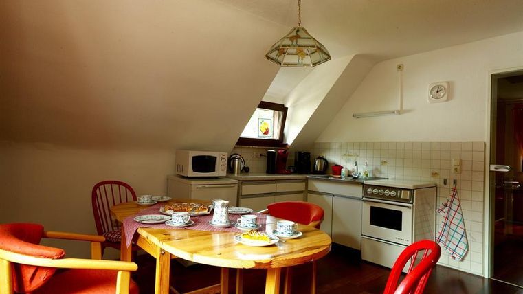 Eine gemütliche Küche mit einem runden Holzertisch und roten Stühlen. Im Hintergrund sind Küchengeräte und ein Fenster, das natürliches Licht hereinlässt.