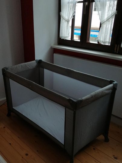 Ein schlichtes Reisebett aus grauem Material steht in einem Raum. Es hat eine flache Matratze und ist von Fenstern umgeben.