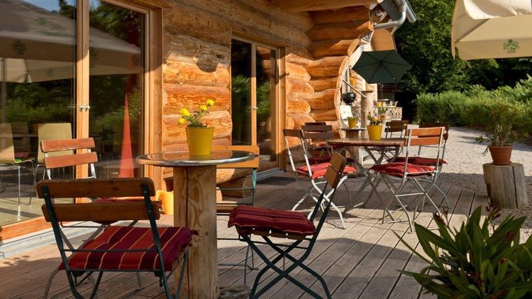 Eine gemütliche Terrasse mit Holzstühlen und einem Tisch. Im Hintergrund sind grüne Pflanzen und ein Holzhaus sichtbar.