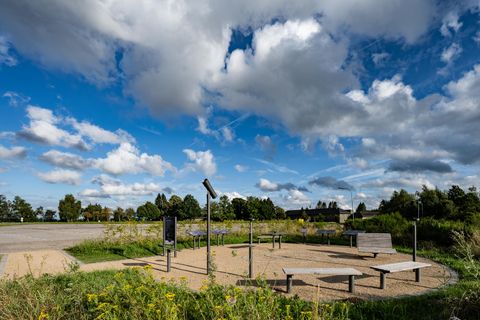 Ein ruhiger Platz im Freien mit Bank und Informationsschildern. Der Himmel ist blau mit weißen Wolken und im Hintergrund sind Bäume sichtbar.