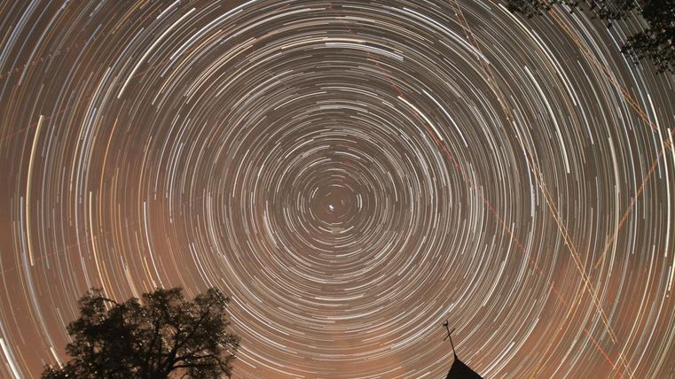 Ein faszinierender Sternenhimmel mit spiralförmigen Sternenspuren. Im Vordergrund sind Silhouetten von Bäumen und einem Dach sichtbar.