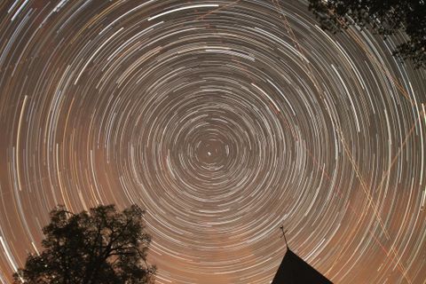 Das ild zeigt den Sternenhimmel bei NAcht über Wollseifen. Durch langzeitbelichtung sieht es so aus als würden die Sterne Kreise um dem Kirchturm ziehen.