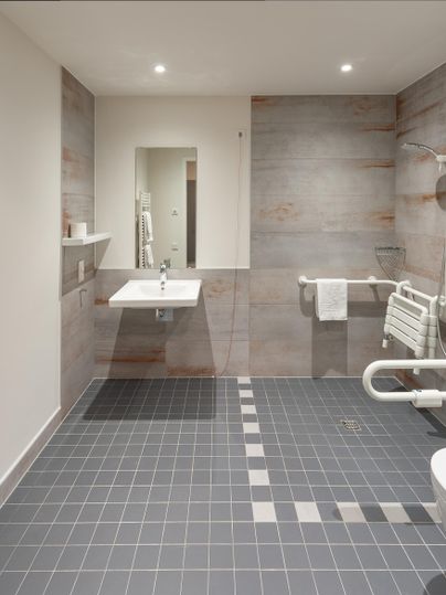 Ein modernes Badezimmer mit grauen Fliesen und einem Waschbecken. Es gibt Haltegriffe und eine begehbare Dusche für Barrierefreiheit.