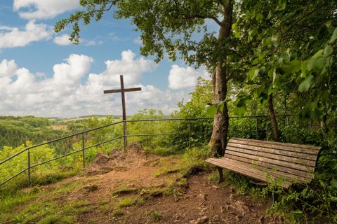 Eine Holzbank steht an einem Aussichtspunkt mit einem Kreuz und Blick auf die grüne Landschaft. Die Wolken am blauen Himmel verleihen der Szene ein friedliches Ambiente.