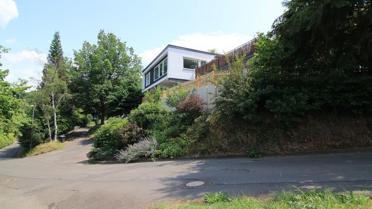 Ein modernes Haus auf einem Hügel, umgeben von üppigem Grün. Die Straße windet sich sanft um das Grundstück.