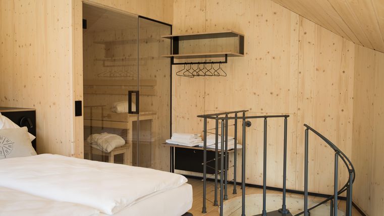 Ein modernes Zimmer mit Holzwänden und einem gemütlichen Bett. Eine Wendeltreppe führt zu einem weiteren Bereich, und es gibt eine Glaswand zur Dusche.