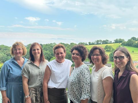 Eine Gruppe von sechs Frauen steht vor einer malerischen Landschaft mit sanften Hügeln. Sie lachen und wirken freundlich, während sie zusammen posieren.
